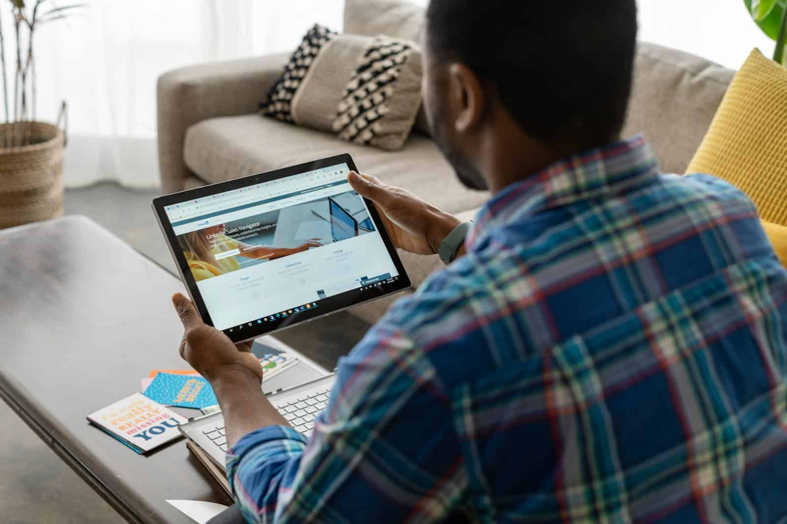 LinkedIn Marketing, Man with plaid shirt browsing internet on tablet at home, seated on sofa.